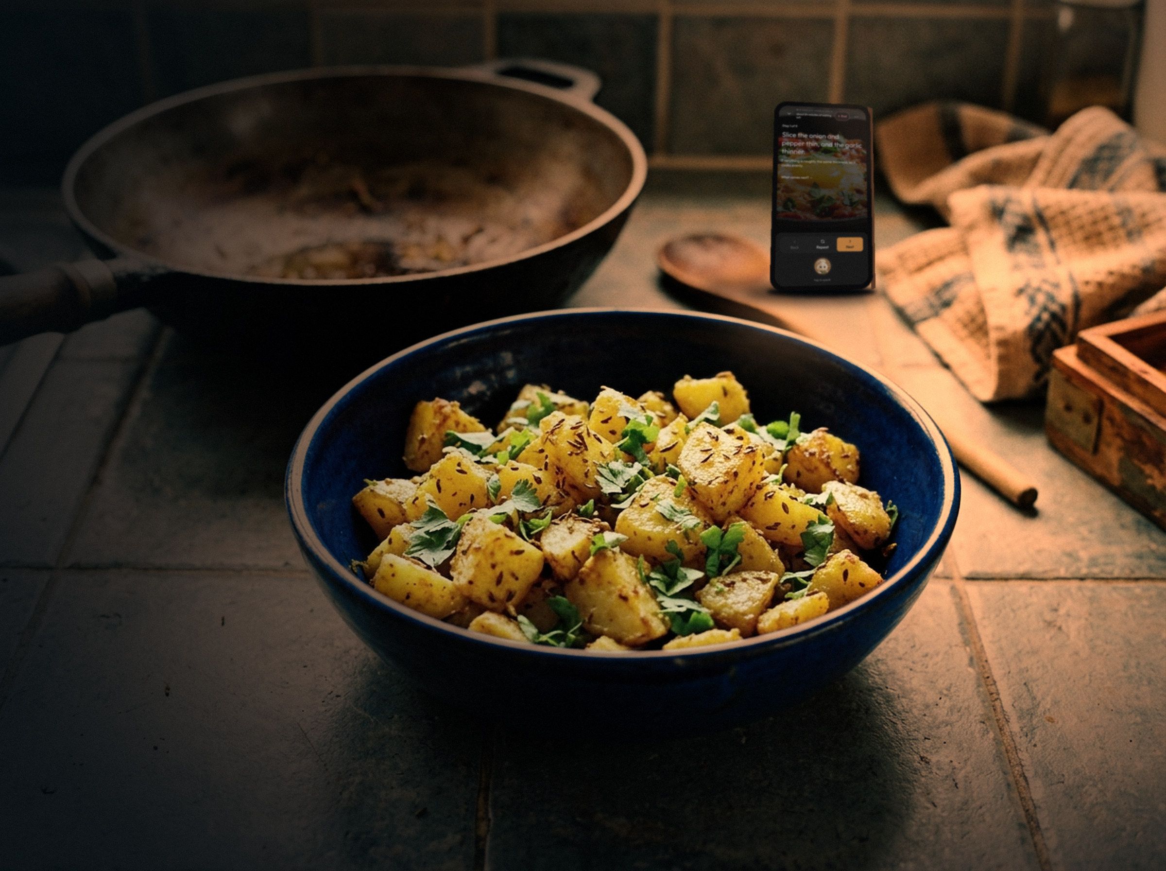 A phone propped at the back of a kitchen counter at night, beyond a pan and a bowl of jeera aloo, showing Waft’s cooking screen: step 1 of 9 in large type, with back, repeat and next buttons.
